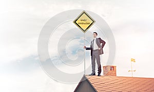 Young businessman on house brick roof holding yellow signboard.