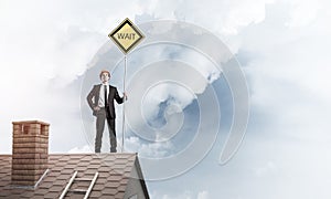 Young businessman on house brick roof holding yellow signboard.