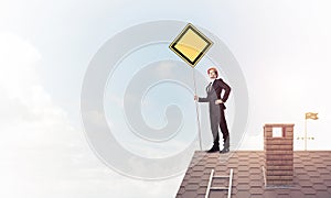 Young businessman on house brick roof holding yellow signboard.