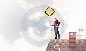 Young businessman on house brick roof holding yellow signboard.