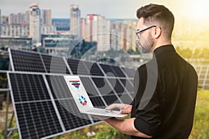 Young businessman with laptop near solar panels and beautiful view of cityscape. Alternative energy source