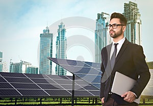 Young businessman with laptop near solar panels and beautiful view of cityscape. Alternative energy source