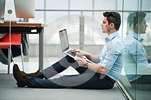 A young businessman with laptop sitting on the floor in office, working.