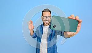 Young businessman laughing and waving while video conferencing on smart phone over blue background