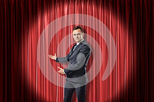 Young businessman lit up by limelight making a presenting gesture against red theater curtain.