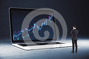 Young businessman looking at huge laptop with forex chart on screen. Dark background. Trade, finance and money concept