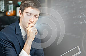Young businessman looks seriously working with tablet, wear suit, sitting in hotel cafe with copy space for background. Young man