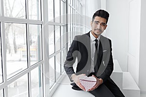 Young businessman in love sitting on windowsill with gift in form of heart.