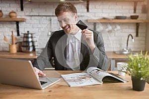 Young businessman making online payment with bank card in laptop in the kitchen while drinking morning coffee before going to