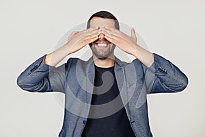 Young businessman man with a beard in a jacket covering his eyes with his hands smiling cheerful and funny. Blind concept.