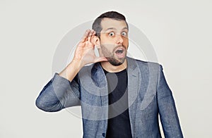 Young businessman man with a beard in a jacket, smiling with his hand above his ear, listening to rumors or gossip. Deafness