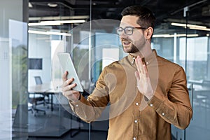 A young businessman man is standing in the office center, holding a tablet in his hands and talking on a video call