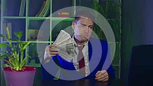 Young businessman with money in modern workplace. Serious businessperson waves money as fan, sitting in office.