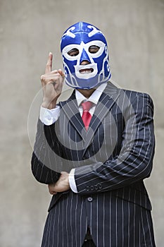 Young businessman in pinstripes suit and wrestling mask pointing up over gray background