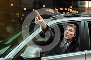 Young businessman pointing with his finger from the open car window, looks amazed, big city life