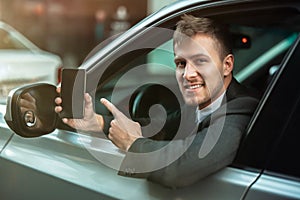 Young businessman pointing with his finger on his smartphone from the open car window, looks happy, big city life