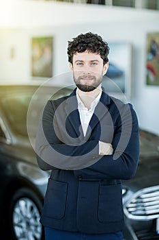 Young businessman or salesman with hands crossed in front of car in showroom