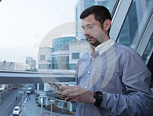 Young businessman with a serious face is browsing the news on a mobile phone against the background of a paned window on a high f