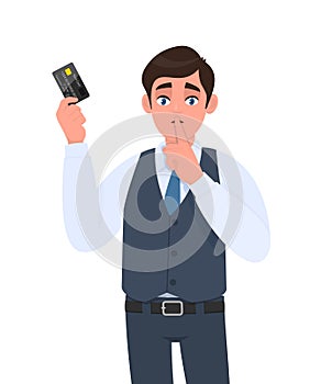 Young businessman showing credit, debit card. Man asking silence and gesturing finger on lips. Sh, shh, keep quiet. Person in vest