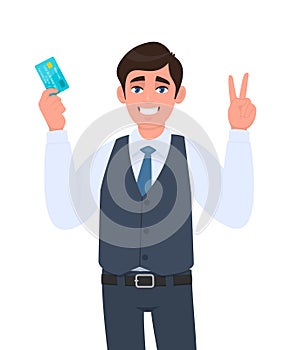 Young businessman showing credit, debit card. Man making or gesturing victory, V, peace or two sign. Trendy person in waistcoat.