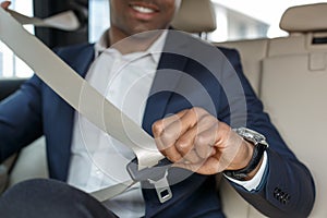 Young businessman sitting on back seat in car fastening belt close-up blurred background cheerful