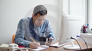 Young businessman sitting by the computer in stylish modern office and taking notes using his pencil and phone. Computer