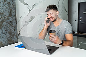 Young businessman sitting indoors at home at the kitchen and talking by mobile phone while using laptop computer