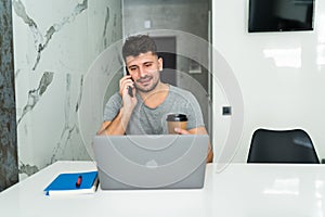 Young businessman sitting indoors at home at the kitchen and talking by mobile phone while using laptop computer
