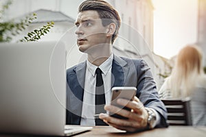 Young businessman sitting next to laptop and holding mobile phone in hand.