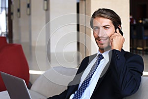 Young businessman sitting relaxed on sofa at hotel lobby making a phone call, waiting for someone.