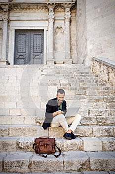 Young businessman sitting on steps of city building and using his phone