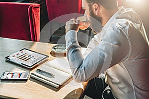 Young businessman is sitting at table,drinking coffee.On table is tablet computer,smartphone with graphs