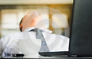 Young businessman is sleeping with a laptop and a coffee cup on the desk. Businessman struggling with drowsiness at workplace