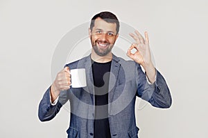 Young businessman with a smile, a man with a beard in a jacket, holding a cup of coffee and showing the gesture everything is ok,