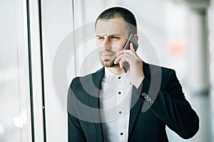Young businessman speaking on mobile phone looking away in office standing beside window