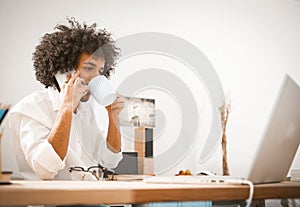 Young businessman speaks on mobile phone while working with a computer. Arab guy drinks coffee while sitting at a desk