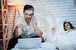 A young businessman speaks on the phone and works behind laptop in bed at home.