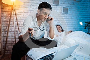 A young businessman speaks on the phone and works behind laptop in bed at home.