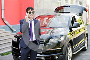 Young businessman in suit and sunglasses talking on phone next to expensive car, outdoors.