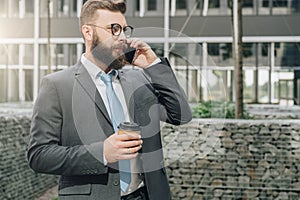 Young businessman in suit and tie is standing outdoor, drinking coffee and talking on his cell phone.Man is working.