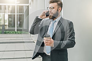 Young businessman in suit and tie is standing outdoor, drinking coffee and talking on his cell phone.Man is working.