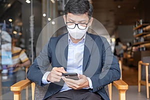 Young Businessman in suit wearing surgical face mask and using smartphone, man typing touchscreen mobile phone in office or cafe.