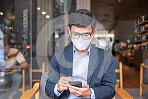 Young Businessman in suit wearing surgical face mask and using smartphone, man typing touchscreen mobile phone in office or cafe.