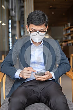 Young Businessman in suit wearing surgical face mask and using smartphone, man typing touchscreen mobile phone in office or cafe.