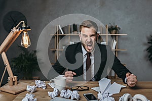 Young businessman in suit yelling while sitting at messy workplace