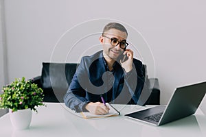 Young businessman talking on cell phone and using laptop in office