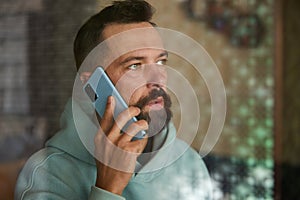 Young businessman talking on his phone indoor through glass. Bearded man in casual clothes using his smartphone.