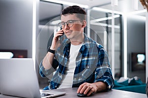 Young businessman talking on phone sitting at kitchen table, busy entrepreneur working distantly from home with laptop