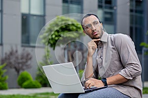 Young businessman thinking sitting outside office building with laptop, man serious and concentrated solving current