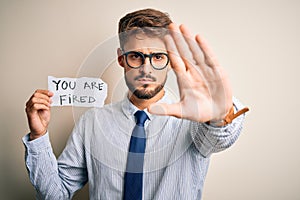 Young businessman wearing glasses holding paper with you are fired message with open hand doing stop sign with serious and